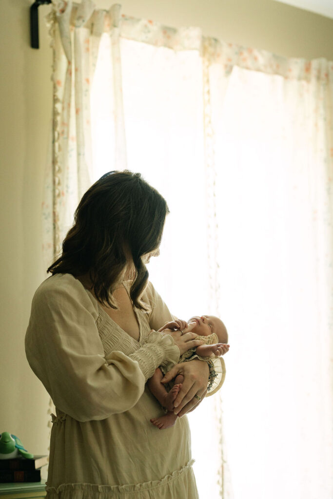 Mother standing beside a bright window holding her newborn
